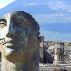 Pompeje: miasto grzechu. Monumentalna głowa rzeźby na tle ruin i Wezuwiusza.