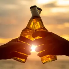 Dwie ręce wznoszą butelki piwa do toastu o zachodzie słońca. W Niemczech od ilu lat można pić alkohol? Od 16 roku życia.