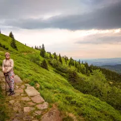 Kobieta na kamienistej ścieżce, podziwia widok na zielone zbocze górskie. To żółty szlak na Śnieżkę, pełen drzew i chmur na niebie.