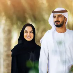 Uśmiechnięta kobieta w tradycyjnym stroju arabskim damskim i mężczyzna w białej dżalabiji spacerują wśród palm.