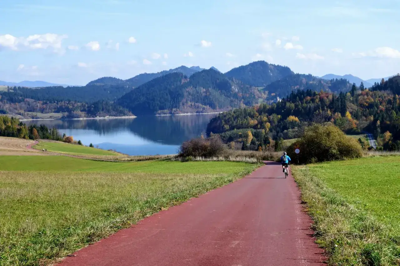 Rowerzysta na czerwonej ścieżce rowerowej Velo Dunajec, otoczonej zielonymi polami i malowniczym jeziorem, w tle góry.